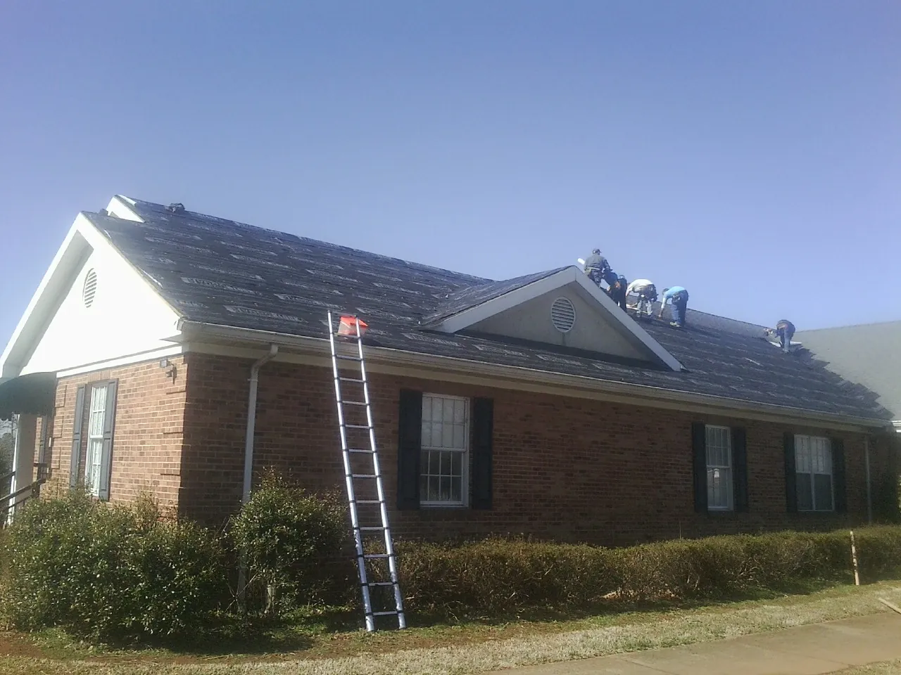 Crew tearing shingles off a brick ranch roof with a ladder set — Emergency Roof Repair work by our crew in Ashuelot