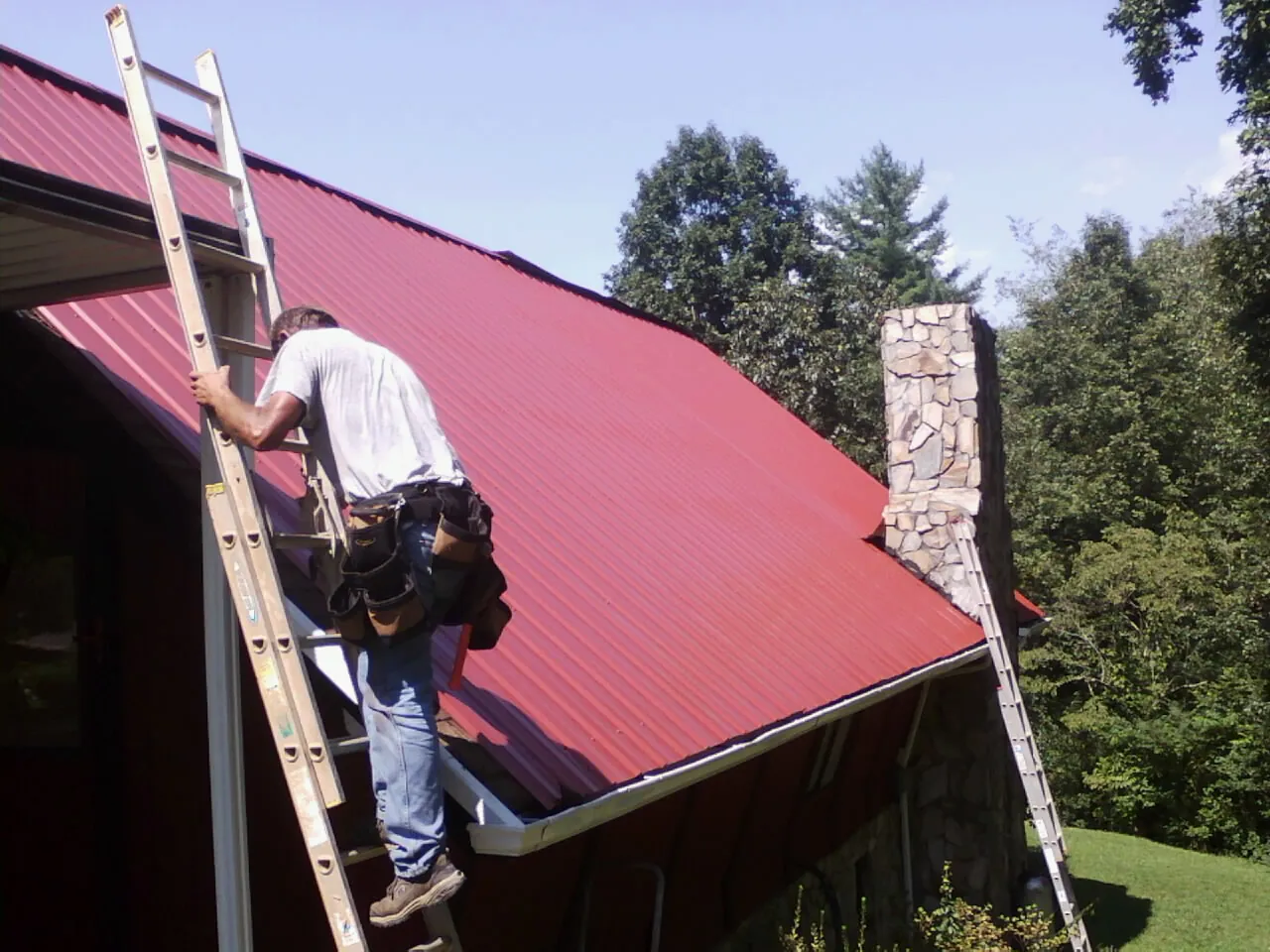 Roofer on a ladder beside a red metal roof and stone chimney — Soffit & Fascia Repair by Roofing Brothers in Ashuelot
